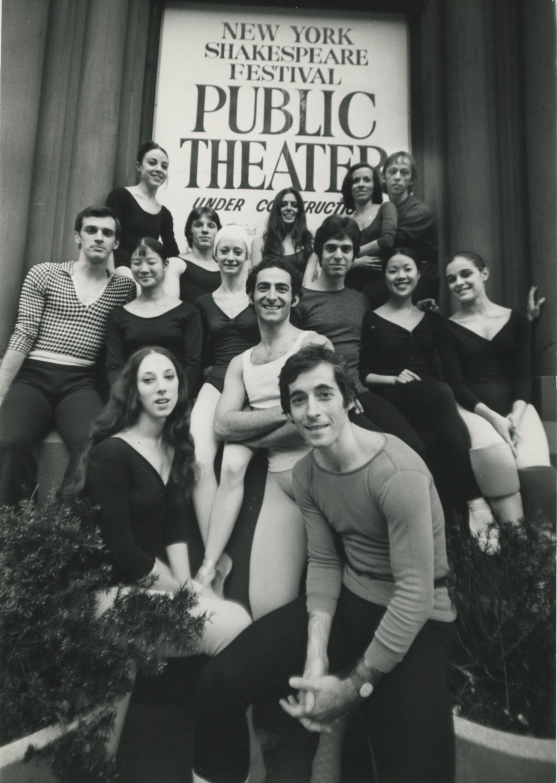 A black and white photo of Eliot Feld and his company dancers, posing together in front of a sign that reads &quot;New York Shakespeare Festival Public Theater.&quot;
