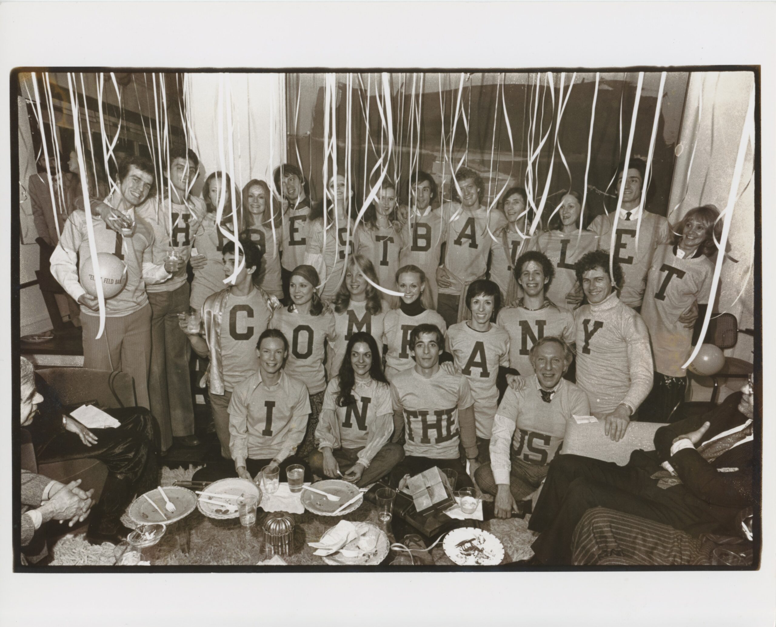 A black and white photo of Eliot Feld and his company dancers. They wear t-shirts with block lettering that spell out &quot;The Best Ballet Company in the US.&quot;