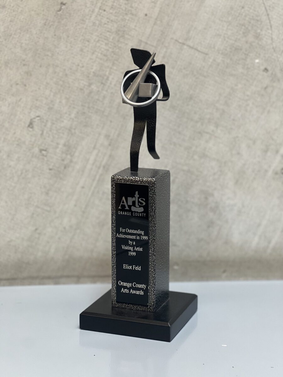 Close up of a trophy with an abstract stick figure at the top. The engraved text reads &quot;Arts Orange County/ For Outstanding Achievement in 1999 by a Visiting Artist 1999/ Eliot Feld/ Orange Country Arts Awards.&quot;