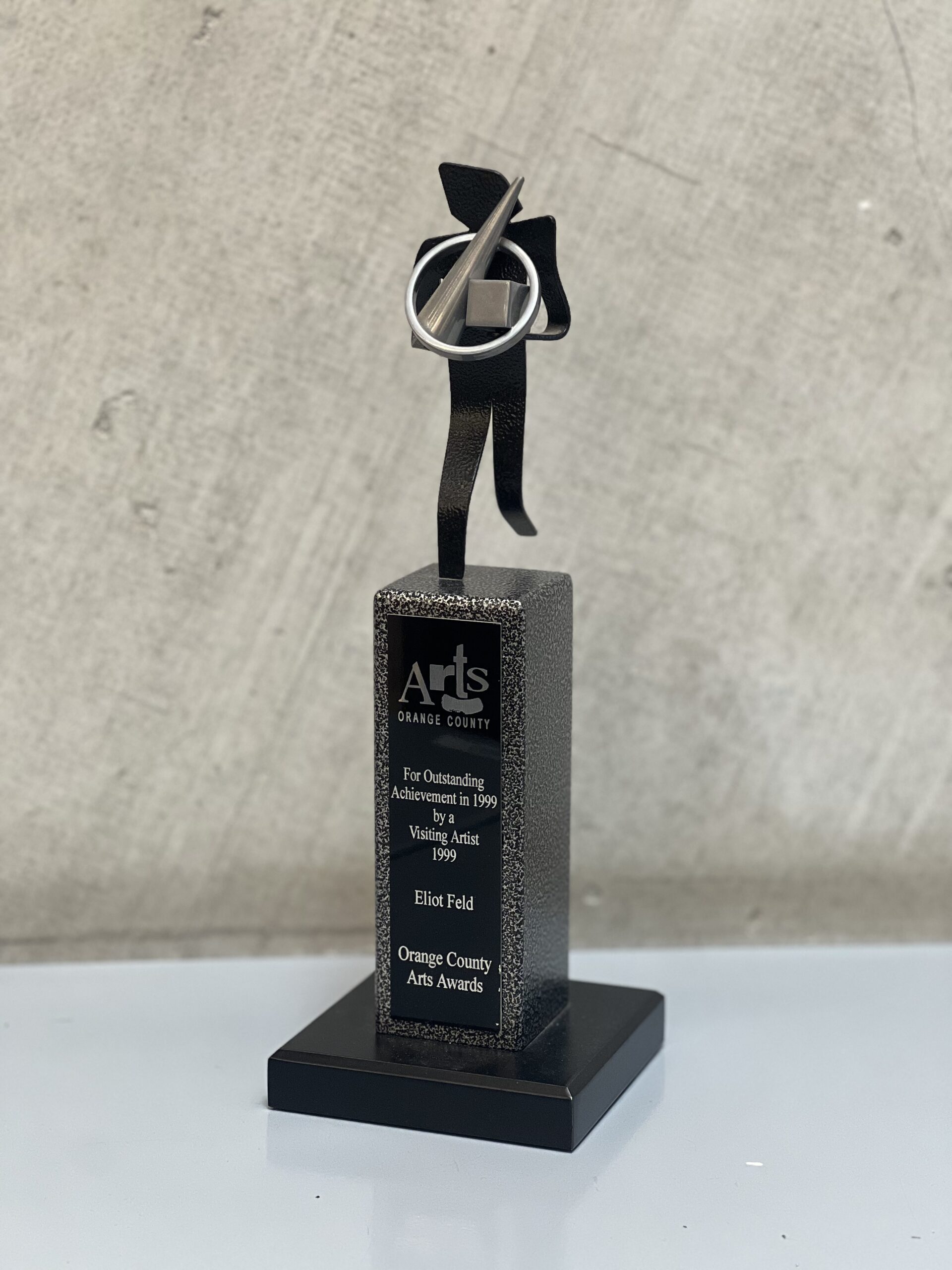 Close up of a trophy with an abstract stick figure at the top. The engraved text reads &quot;Arts Orange County/ For Outstanding Achievement in 1999 by a Visiting Artist 1999/ Eliot Feld/ Orange Country Arts Awards.&quot;
