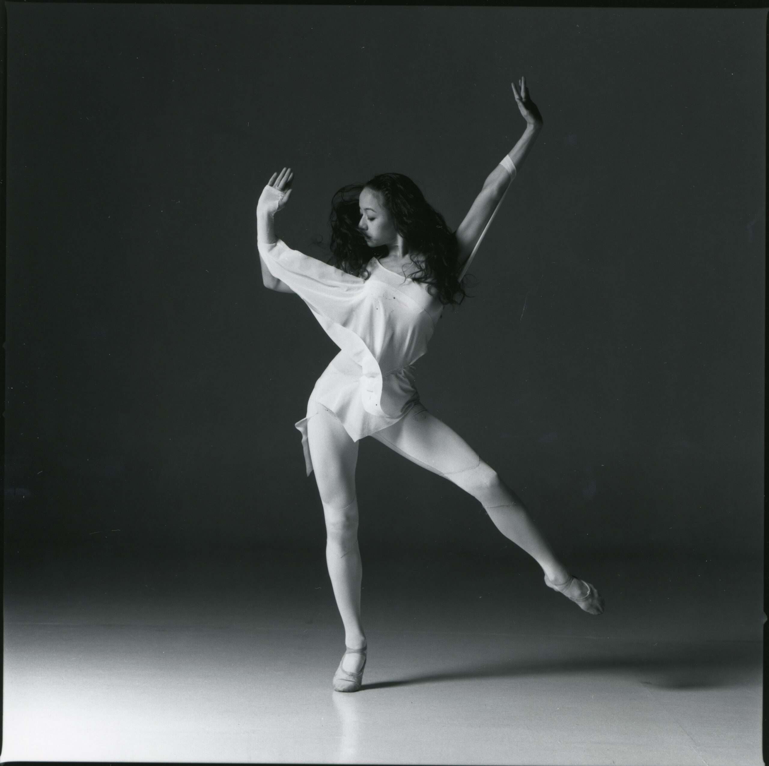 Black and white photo of dancer Ha Chi Yu, dancing in a silky white top and ballet shoes. She balances on one leg.