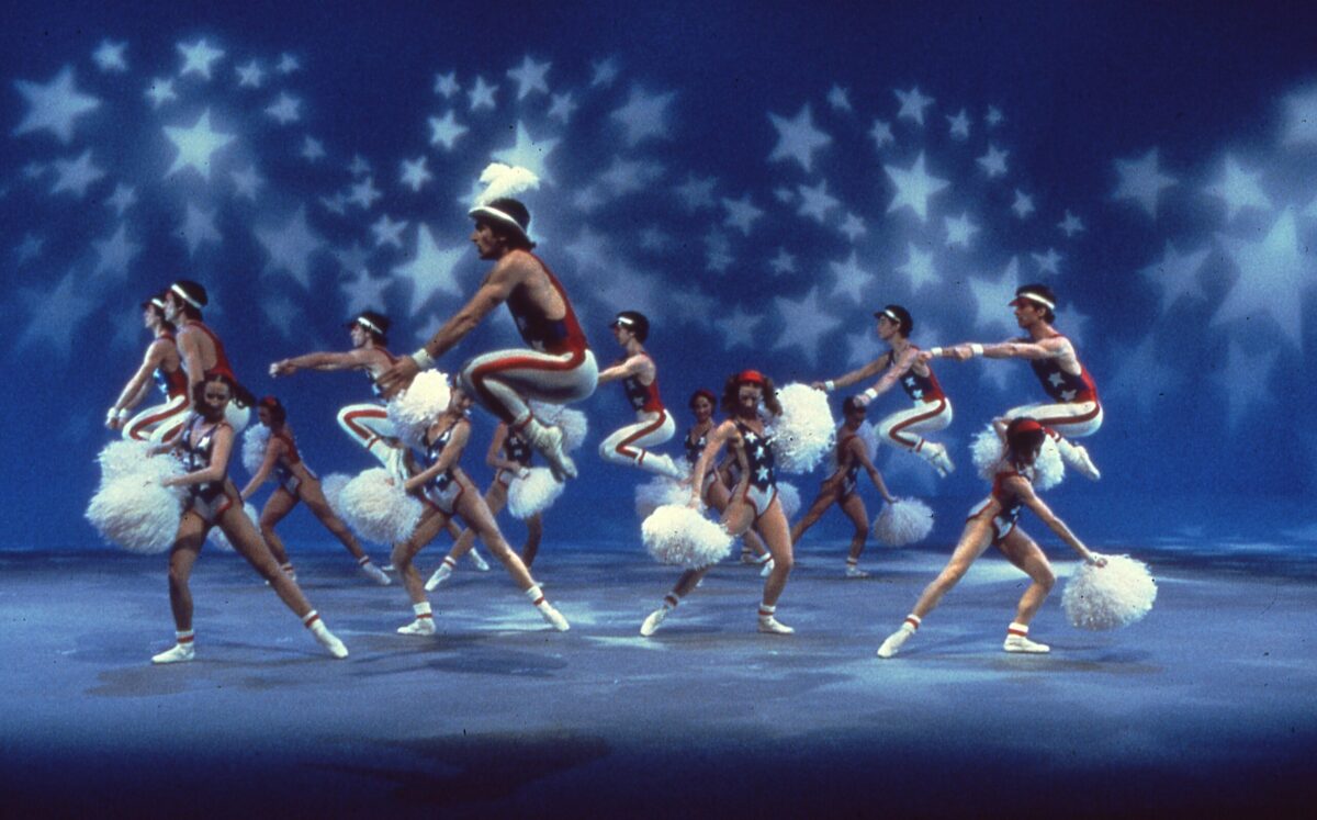 Dancers in red, white, and blue costumes with stars and stripes hold white pom-poms while performing Eliot Feld&#039;s &quot;Half Time.&quot; Stars are projected onto the scrim behind the dancers.