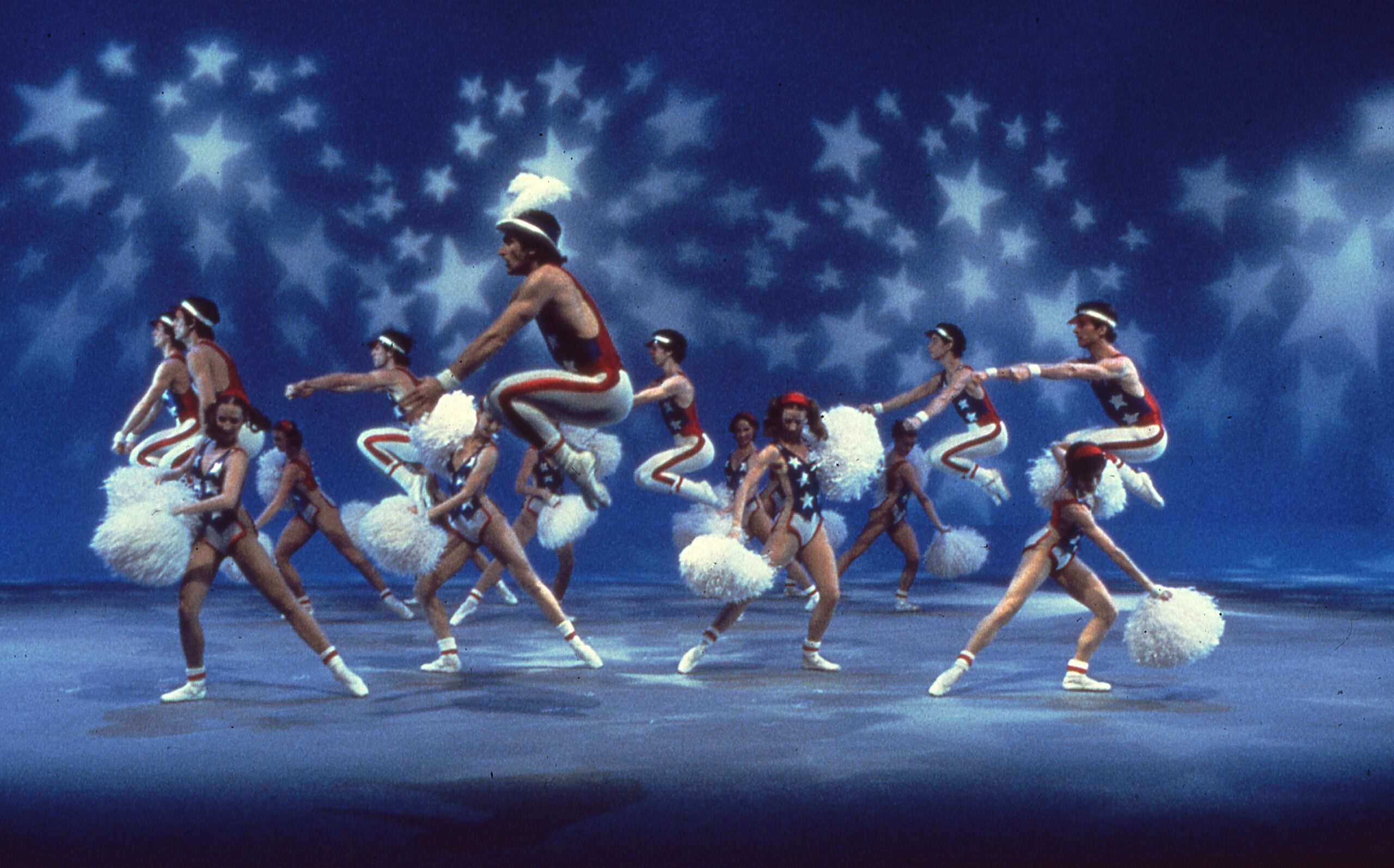 Dancers in red, white, and blue costumes with stars and stripes hold white pom-poms while performing Eliot Feld&#039;s &quot;Half Time.&quot; Stars are projected onto the scrim behind the dancers.