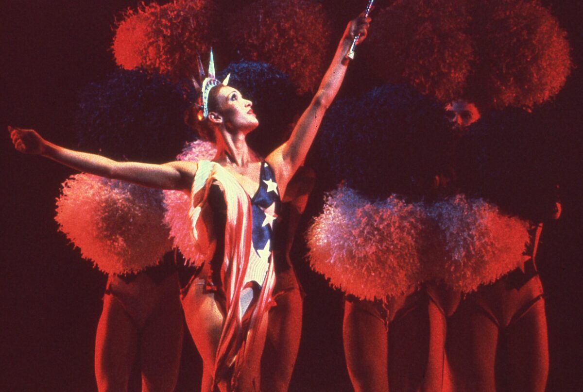 A dancer wearing a red, white, and blue leotard with stars and a Statue of Liberty crown performs Eliot Feld&#039;s &quot;Half Time&quot; on stage. Four dancers stand in a line behind her, holding red, white, and blue pom-poms that cover their faces.