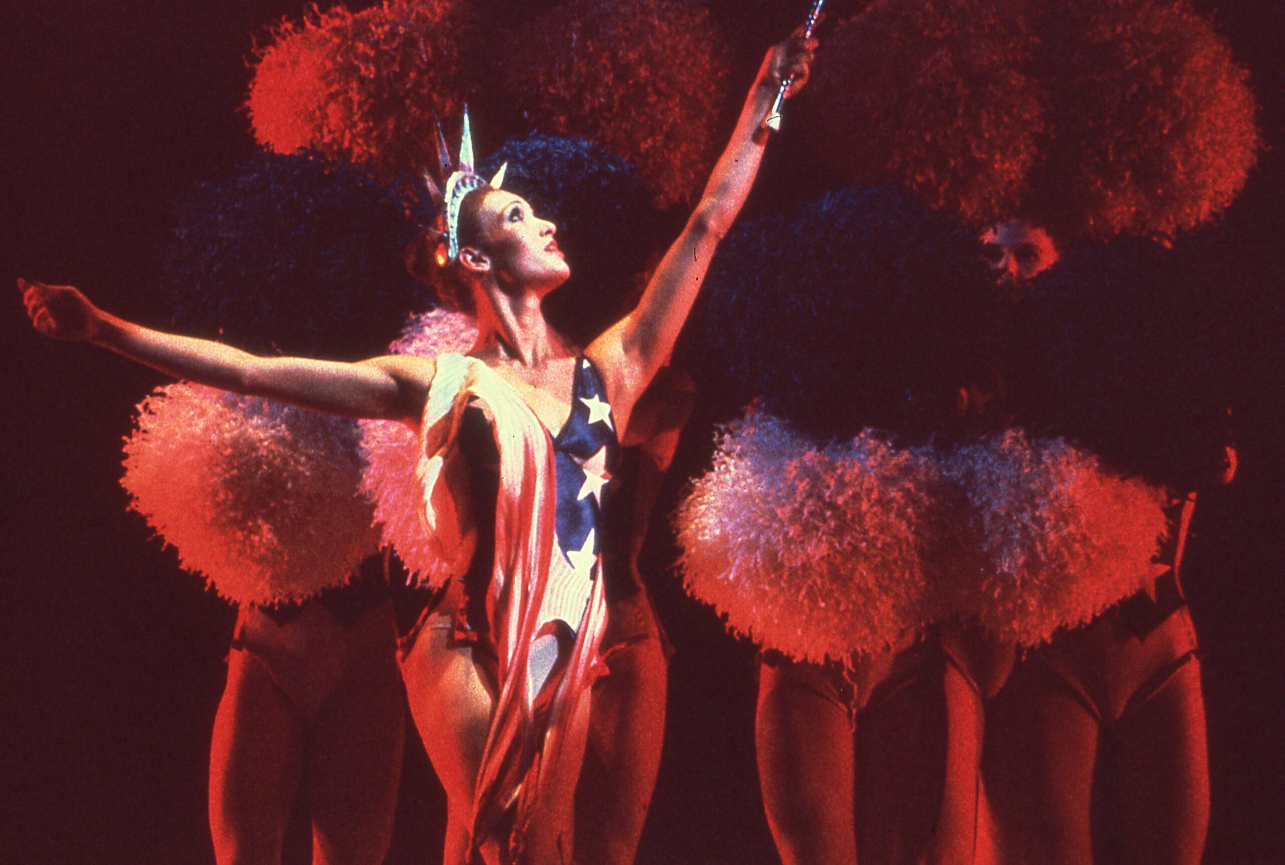 A dancer wearing a red, white, and blue leotard with stars and a Statue of Liberty crown performs Eliot Feld&#039;s &quot;Half Time&quot; on stage. Four dancers stand in a line behind her, holding red, white, and blue pom-poms that cover their faces.