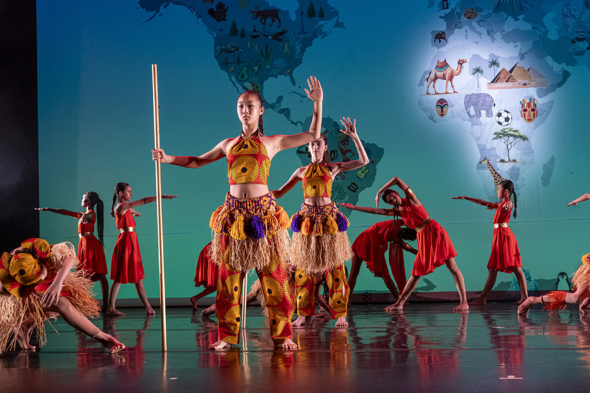 Ballet Tech students perform a West African piece on stage, standing in various formations. Some dancers wear raffia skirts with colorful patterned fabrics, while others wear orange tunics with gold waistbands.