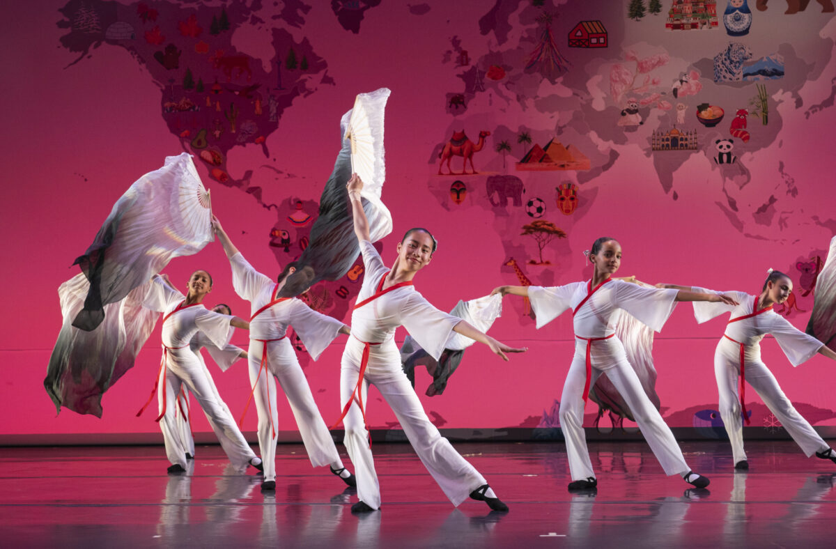 An ensemble of young dancers on stage wear white robes with red ties, inspired by traditional Chinese folkdance attire. They dance in unison waving large fans that billow behind them. The backdrop features a world map with various cultural icons and images.