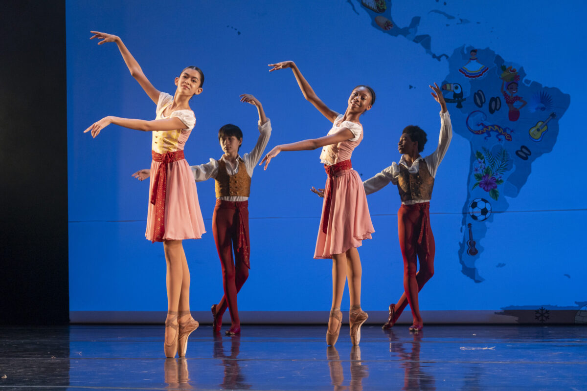Four dancers perform a ballet piece on stage. Two girls stand on pointe, with arms in fourth position, while two boys stand behind them in b plus. The girls wear corset vests with flowing skirts and red waistbands, while the boys wear red tights with gold vests atop white blouses.