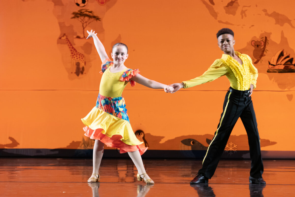 Two young dancers partner in a salsa dance on stage. They hold hands while posing apart, wearing proud expressions. The girl wears a bright yellow dress with colorful accents, and the boy wears a ruffled yellow shirt with black pants.