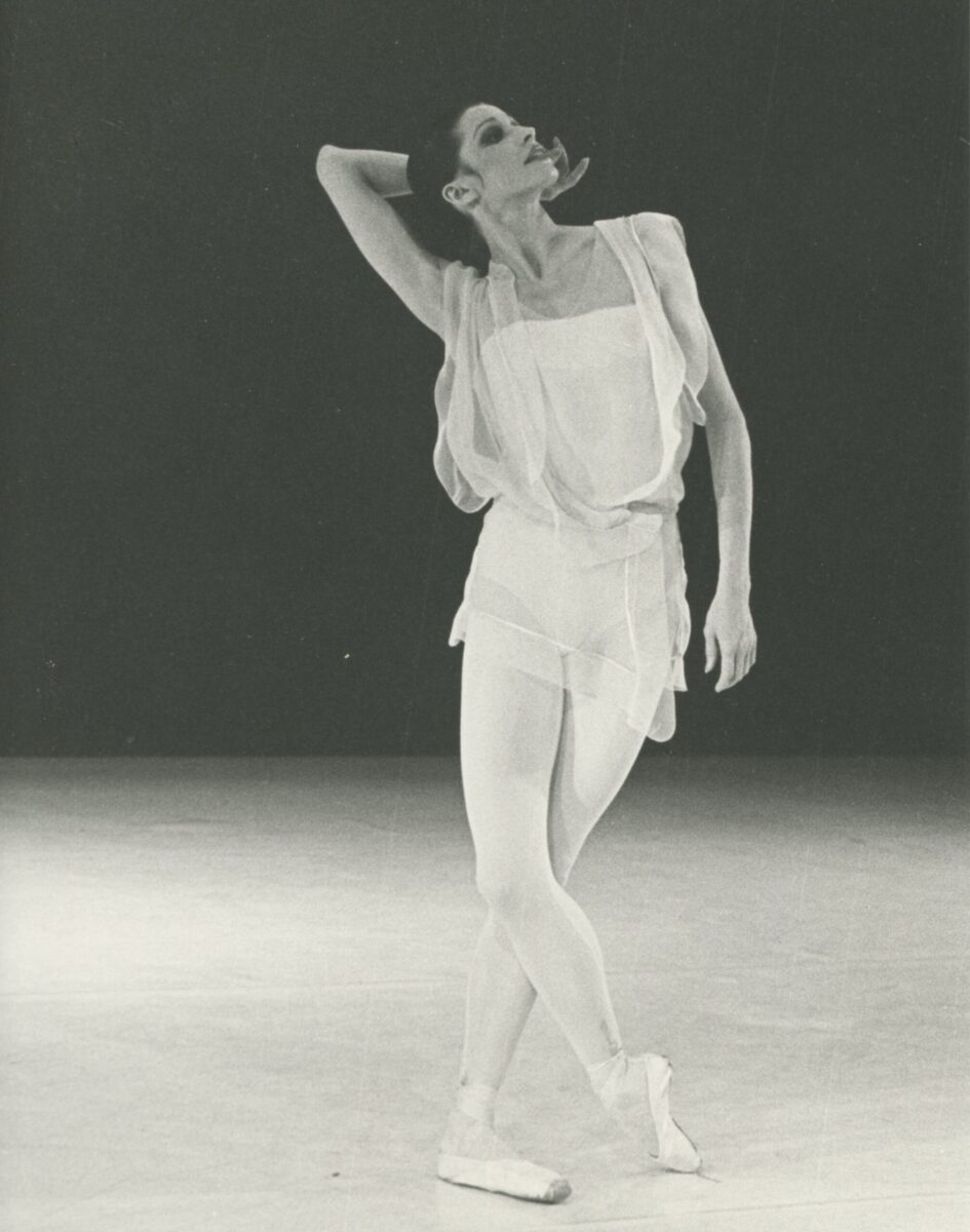 A black and white photo of of dancer Birgit Keil. She stands on stage in pointe shoes, with one leg crossed over the other, her hand gracefully posed behind her head as she gazes upward.