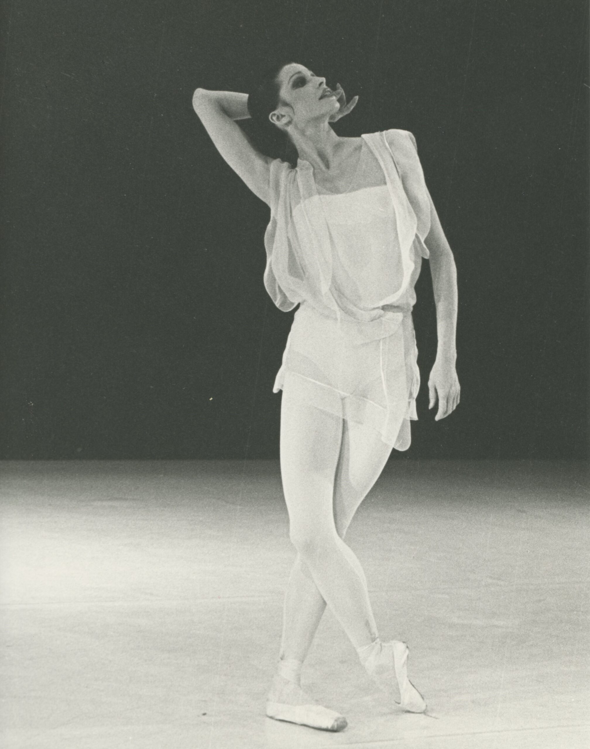 A black and white photo of of dancer Birgit Keil. She stands on stage in pointe shoes, with one leg crossed over the other, her hand gracefully posed behind her head as she gazes upward.