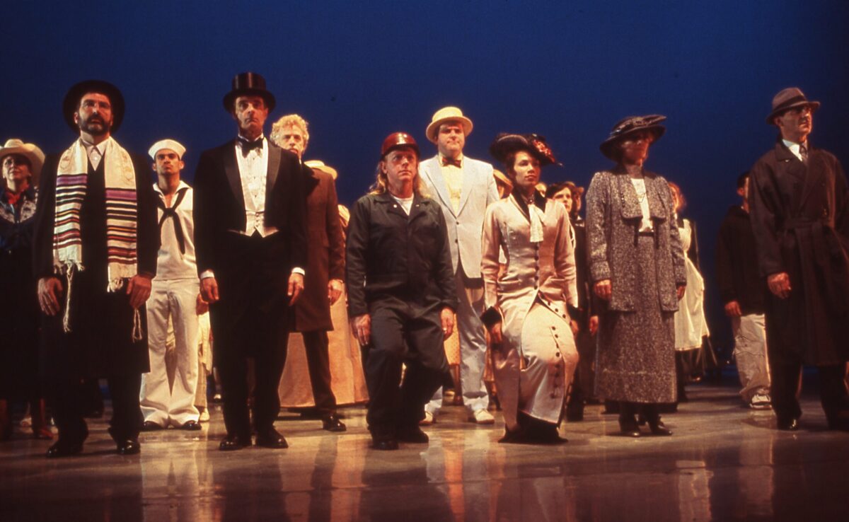 In a performance of Eliot Feld&#039;s &quot;Lincoln Portrait,&quot; a large ensemble of dancers stand on stage, dressed in pedestrian, 1800s America-era clothing.
