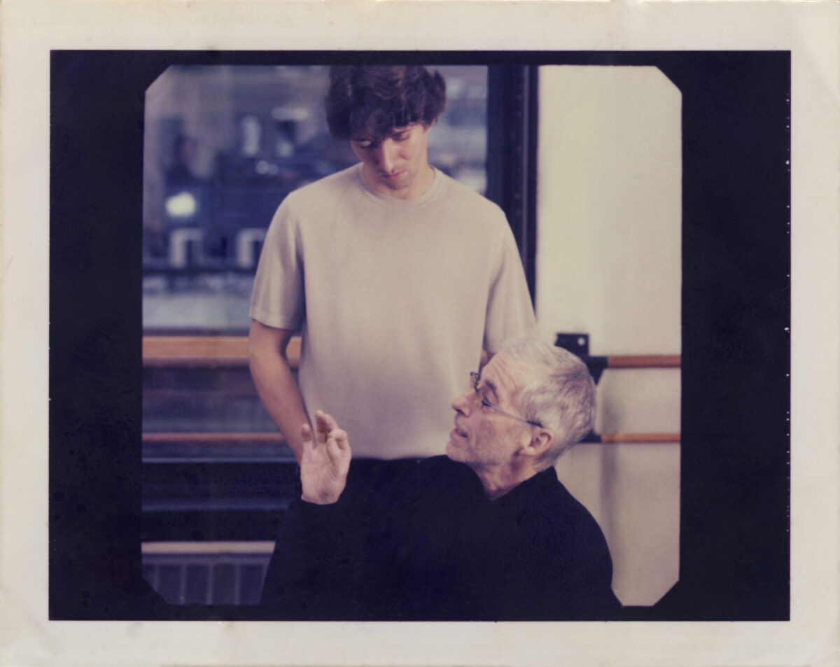 In a dance studio, Damian Woetzel stands behind Eliot Feld, listening as he speaks and gestures with his hand.