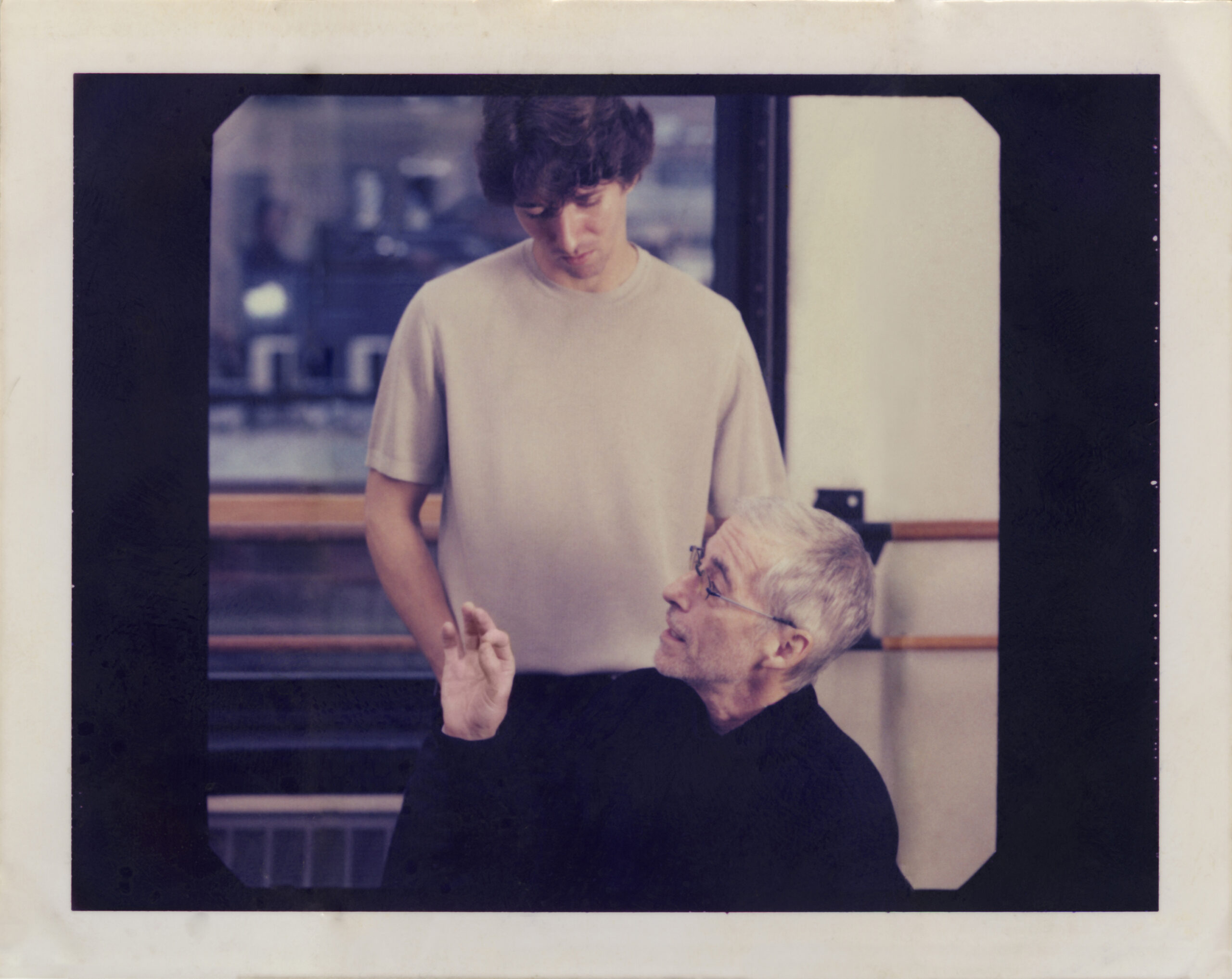 In a dance studio, Damian Woetzel stands behind Eliot Feld, listening as he speaks and gestures with his hand.
