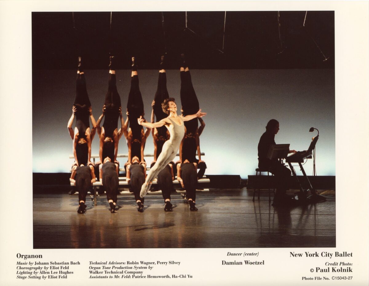 Damian Woetzel jumps gracefully on stage in performance of Eliot Feld's "Organon." An ensemble of dancers poses in an acrobatic formation behind him. A musician in silhouette plays a keyboard to the right of the dancers.