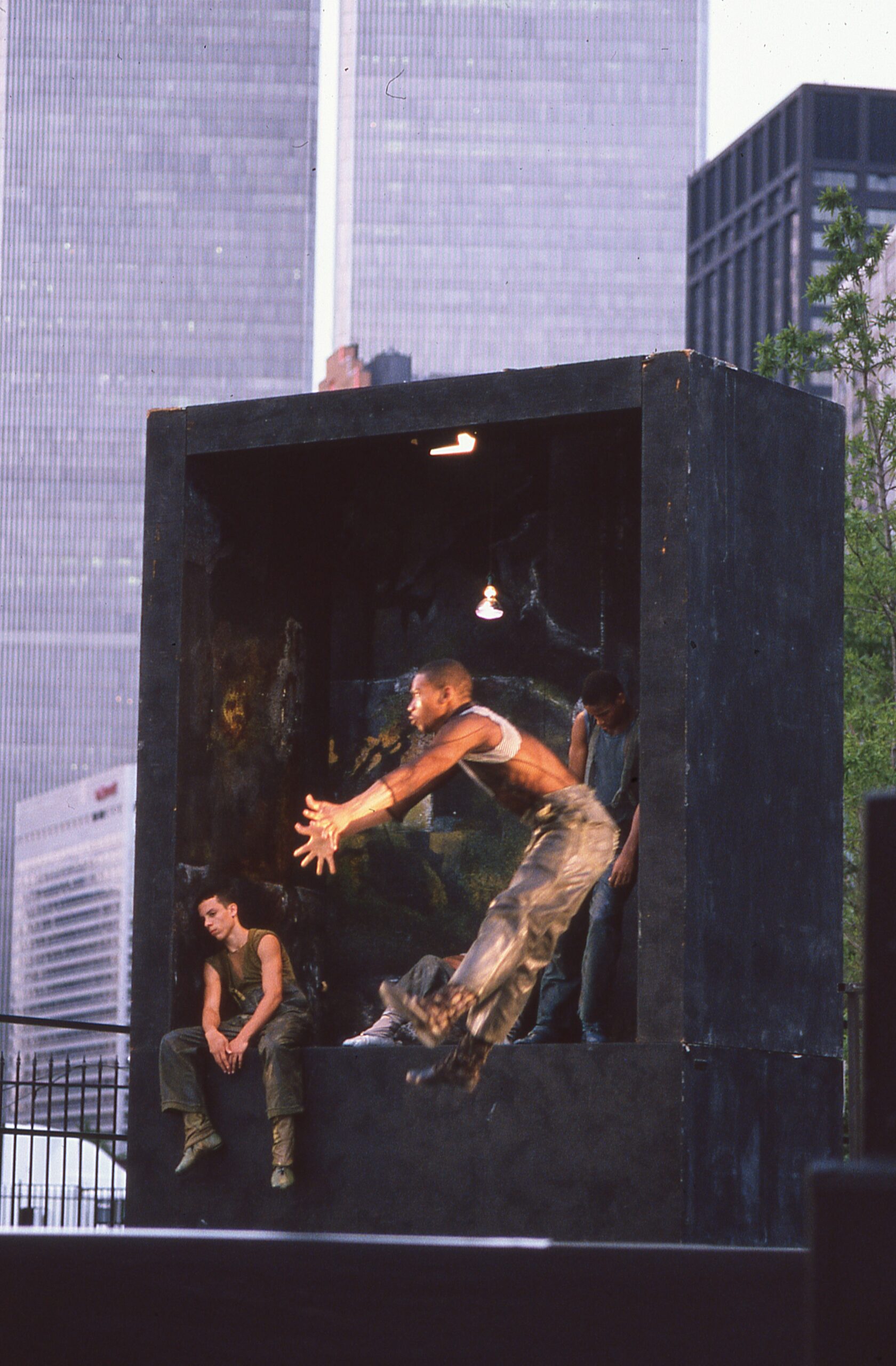Four male dancers perform on an outdoor stage. Three dancers sit and recline atop a cubic platform, and the fourth dancer jumps backwards, while reaching his arms and legs in front of him.