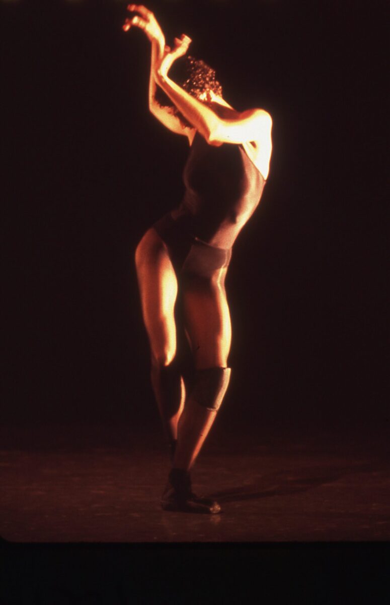 A female dancer on stage wears a leotard and pointe shoes. Standing in 5th position, she sways her hips, head, and arms to the side, covering her face.