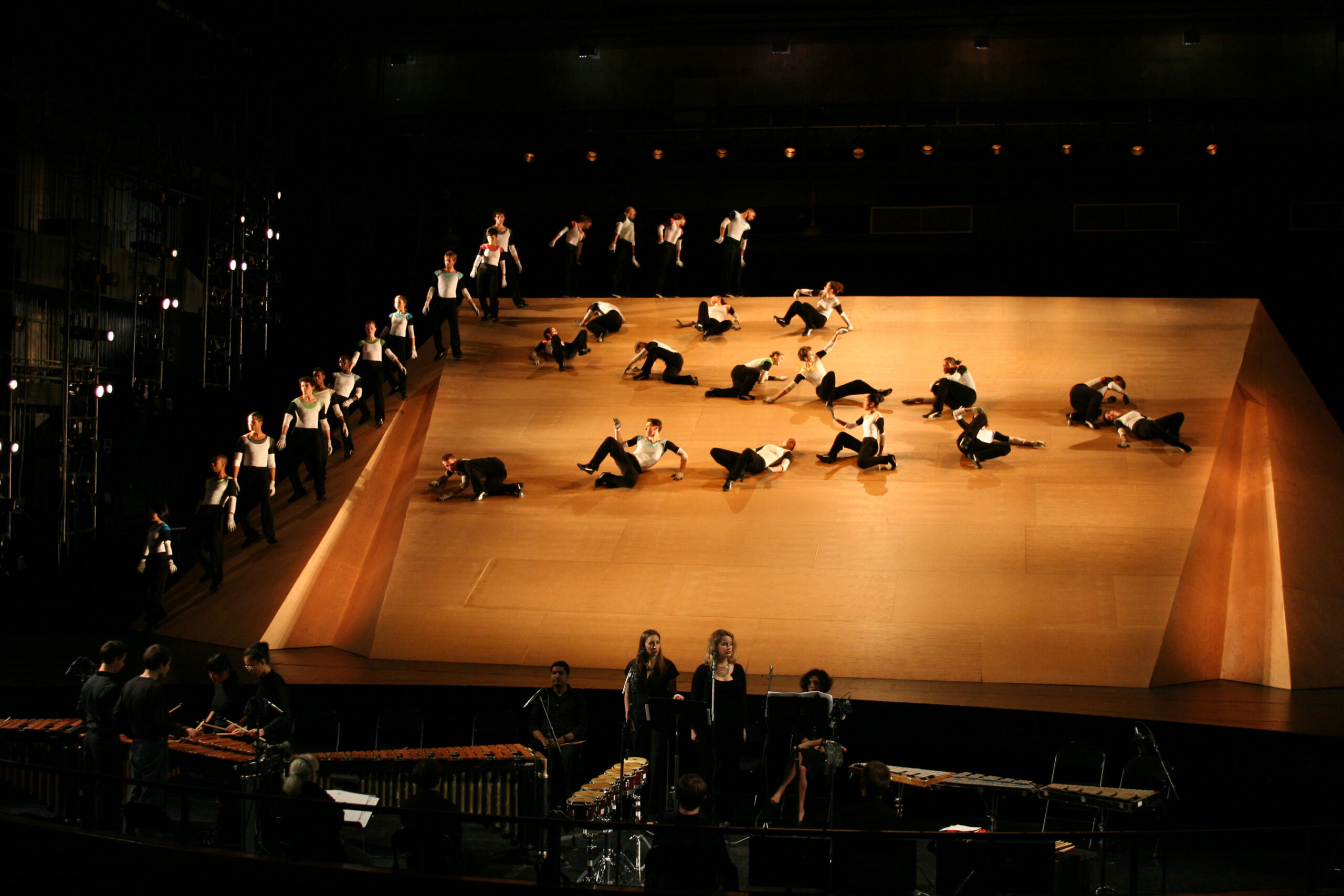 A wide, almost aerial shot of Eliot Feld&#039;s &quot;Sir Isaac&#039;s Apples&quot; performed on stage. One group of dancers stand in a line along part of the perimeter of the stage. They feed into a group of dancers that one by one roll and slide across the stage in a zig-zagging line.