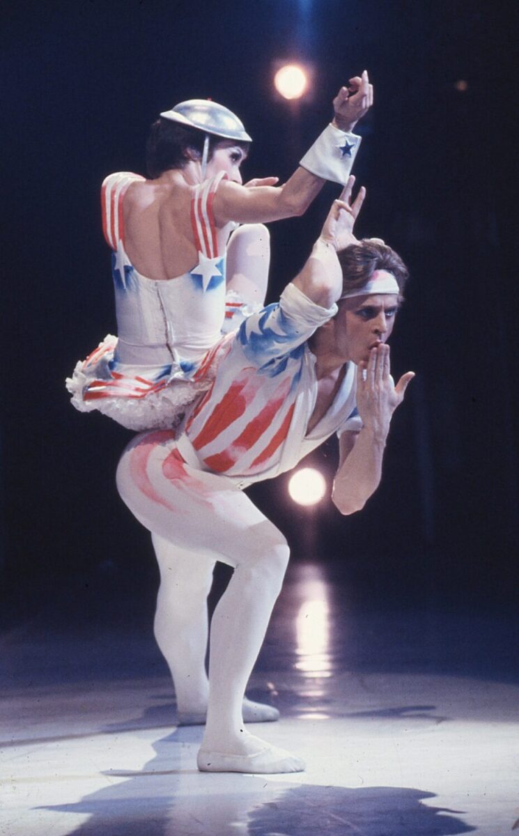 Two dancers performing Eliot Feld&#039;s &quot;Variations on America&quot; partner each other on stage. The woman balances in a seated perch on the mans back. They wear red, white, and blue costumes with stars and stripes, and gesture theatrically towards the audience.