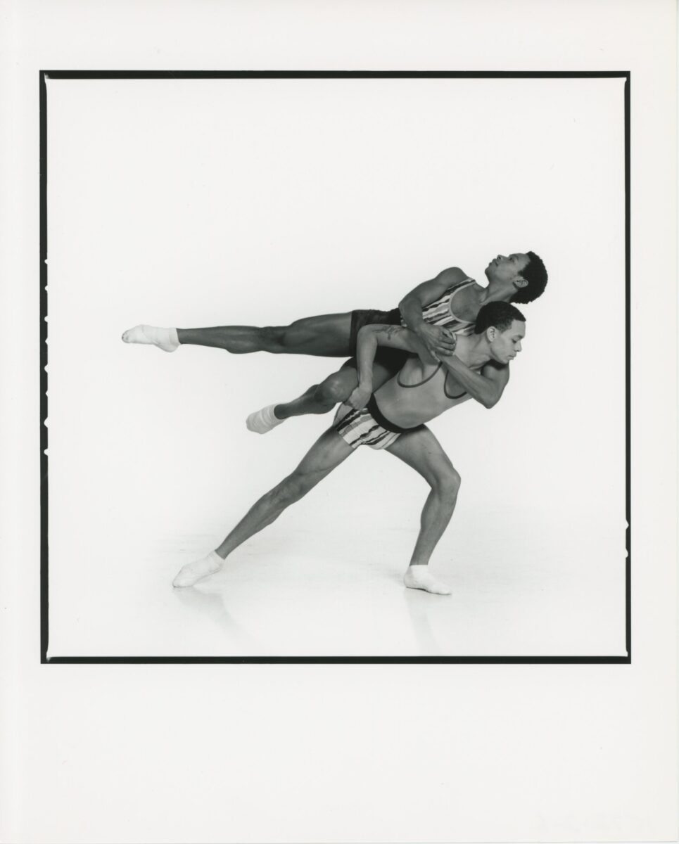 Black and white photo of Jason Jordan and Jassen Virolas partnering each other. Virolas lunges and lifts Jordan on his back. Jordan balances and leans his body sideways, extending his leg to the side, and looking upwards in a graceful pose.