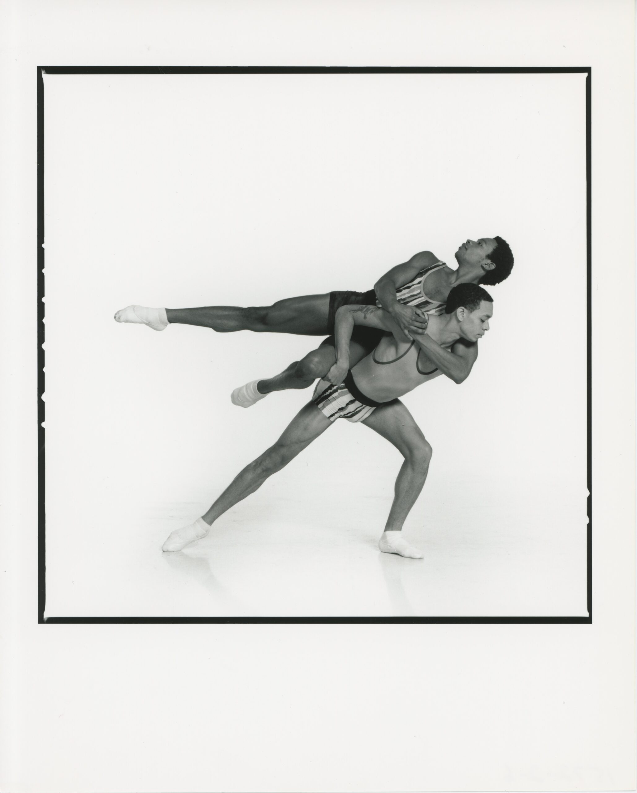 Black and white photo of Jason Jordan and Jassen Virolas partnering each other. Virolas lunges and lifts Jordan on his back. Jordan balances and leans his body sideways, extending his leg to the side, and looking upwards in a graceful pose.