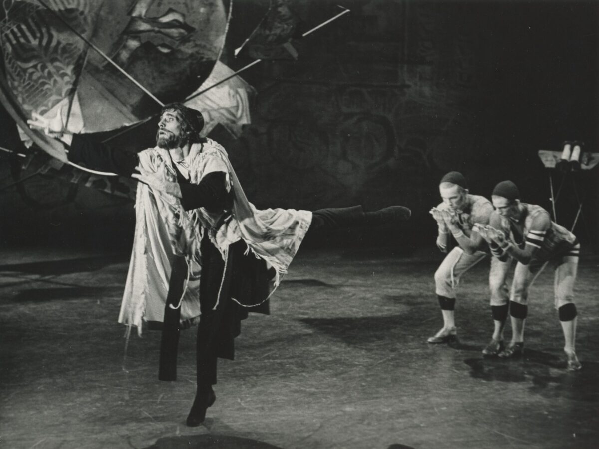 Black and white photo of a performance of Eliot Feld&#039;s &quot;Tzaddik.&quot; Three male dancers perform on stage. One reaches forward in a pique arabesque while the other two crouch behind him, holding their palms in front of their faces.