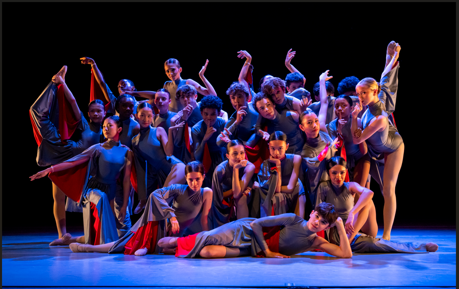 A group of dancers pose in a clump on stage in various dance poses and gestures. They wear gray leotards with red accents, and flowing pants.